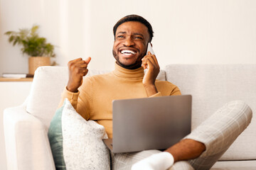 Aufkleber - Good News. Excited African American Man Talking On Cellphone Using Laptop Sitting On Sofa Indoor. Empty Space, Selective Focus
