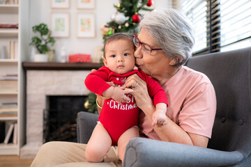 Wall Mural - Grandma is sitting in an armchair, holding her infant grandson, playfully chatting with him in the living room, which is decorated for the upcoming Christmas celebration