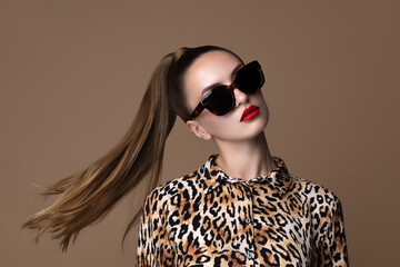 Fashion portrait of a woman in black square sunglasses and leopard print blouse, with sleek high ponytail and red lipstick. Stylish and confident against a beige backdrop. Eyewear glasses accessory