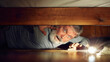 © master1305 - Fearful senior man looking under bed with flashlight, expressing emotions of fear, feel scared. Frightening night at home. Concept of Halloween, human emotions, spooky season