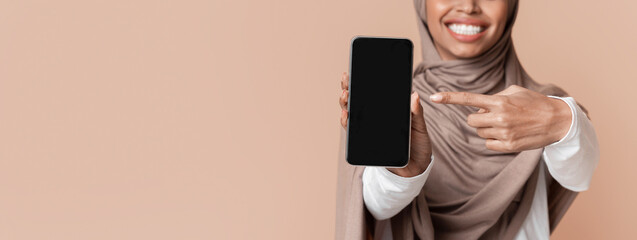 Wall Mural - Afro Muslim Woman In Hijab Pointing At Smartphone With Empty Black Screen In Her Hands, Selective Focus On Device, Yellow Background With Free Space