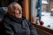 © Milos - A senior man dressed warmly in a thick sweater is sitting comfortably in a cozy room, gazing thoughtfully out the window which shows a snowy landscape and festive decorations.