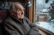 © Milos - An elderly man clad in a brown zipped sweater looks through a window adorned with small Christmas decorations, capturing the serene snowy outdoor landscape and distant house.