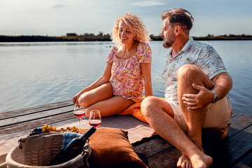 Smiling adult couple sitting by the lake on a pier drinking wine.