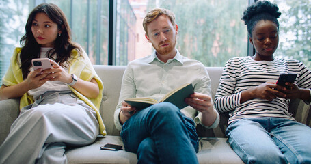 Close up Multi-ethnic group of strangers sitting on grey couch. Man in middle reading book. Girls using mobile devices.