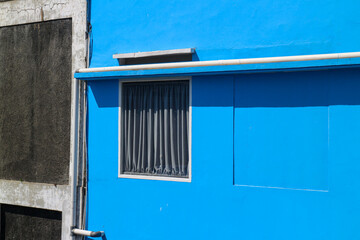 Poster - A blue wall with a window and a gray wall building. A single window with curtains peeks out from a vibrant blue building