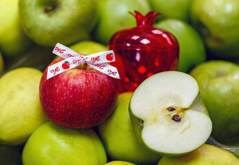 Red apple, a decorative red pomegranate and a half cut apple sit among fresh green apples. Ribbon with the Hebrew inscription words shana tova is attached to it which means Happy New Year. Israel