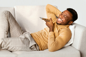 Aufkleber - Positive Black Man Using Mobile Phone Voice Assistant Lying On Couch At Home. Modern Lifestyle