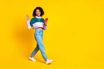 Poster - Full length photo of friendly girl wear striped shirt hold smartphone go empty space waving palm isolated on vivid yellow color background