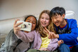 © Connect Images - Three cheerful kids taking a selfie on a couch, with one flashing a peace sign.