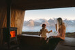 © EVERST - Family mother and child relaxing in cozy mountain hut with fireplace and panoramic window with mountains view, parent traveling with child in Norway autumn vacations trip, mom and kid eating snacks