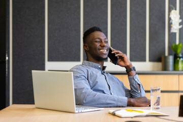 Talking on smartphone, man smiling and working on laptop at office desk