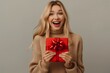 © YURIMA - A happy woman in a cozy sweater holds a gift box with a red bow, showing joy. Possibly celebrating a special occasion, she exudes excitement and delight, radiating happiness and positive emotions