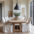 © Chawakorn - A rustic wooden dining table with white linen chairs, a potted plant, and a large pendant light in a farmhouse kitchen.