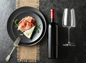 Poster - Bottle of red wine with blue cheese, prosciutto, and rosemary on a black table.