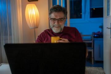 Wall Mural - one man adult caucasian work on laptop at home happy smile with cup of coffee. senior man sitting at desk with laptop computer at home office, looking and smiling at screen