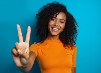 Photo portrait of a pretty young girl showing a v-sign, wearing a trendy orange outfit, isolated against a vibrant blue background. Her bright smile exudes confidence, joy, and positive energy.