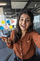 Wall Mural - Office worker smiling and looking at phone camera, businesswoman using smartphone for video call, joyful woman talking with friends and colleagues remotely while sitting at workplace inside office.