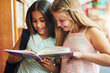 © AK Coop/peopleimages.com - School kids, girls or happy in library with book for educational story, reading fantasy fiction or research. Friends, student or novel at academy for exam assessment, knowledge or learning with smile