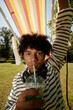 © Seventyfour - Vertical shot of playful African American teenage boy drinking soda with straw looking at camera while kidding around by raising chair in air in summer park, fisheye effect, camera flash