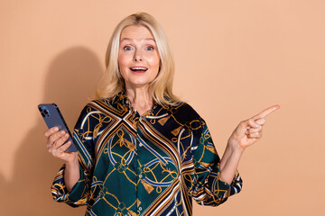 Wall Mural - Photo portrait of blonde senior business woman in stylish shirt using smartphone pointing finger copyspace isolated on beige color background