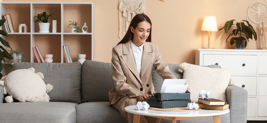 Wall Mural - Beautiful happy female writer with typewriter and books and crumpled paper sitting on sofa at home