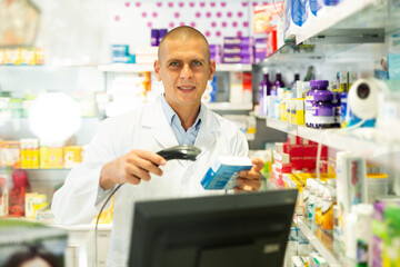 Male pharmacist in lab coat standing at counter in drugstore and using barcode scanner to sale drug package.