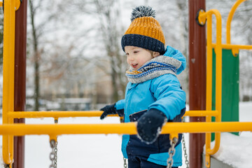 Wall Mural - Adorable little boy having fun on a playground on snowy winter day. Cute child wearing warm clothes playing in a snow.