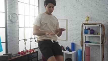 Sticker - A young hispanic man exercises at a gym, performing a balance routine with one leg raised amid various equipment in a well-lit, organized sports center.