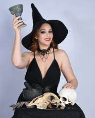 Close up portrait of beautiful  female model wearing black dress, pointy witch hat halloween costume.  Isolated figure on studio background. Holding wine goblet at spooky occult ritual potion table 