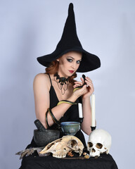 Canvas Print - Close up portrait of beautiful  female model wearing black dress, pointy witch hat halloween costume.  Isolated figure on studio background. Holding snake serpent at spooky occult ritual potion table 