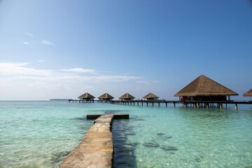 A serene view of overwater bungalows on stilts in a clear turquoise lagoon, with a stone pathway leading towards them. The sky is bright blue with a few clouds, creating a tranquil tropical atmosphere