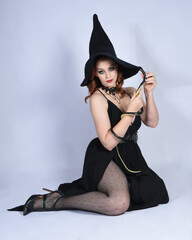 Canvas Print - Full length portrait of beautiful female red head model wearing  glamorous black witch dress with pointy hat halloween costume.  Kneeling pose with gestural arms, isolated figure on studio background