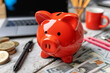 © July P - Red piggy bank on a desk surrounded by coins, bills, a pen, and office supplies. Blurred background. Concept of savings and finance.