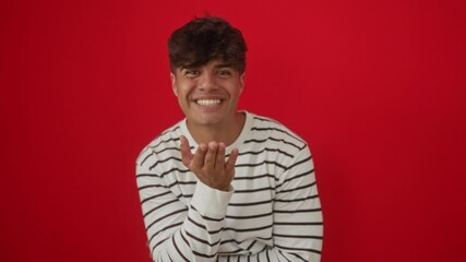 Canvas Print - Young hispanic man wearing stripes t-shirt standing and blowing a kiss with hand in air, showing lovely and sexy expression, over isolated red background, looking at camera with love