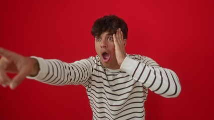 Poster - Pointing young hispanic man wearing stripes t-shirt standing in surprise with open mouth, finger pointing ahead, over isolated red background