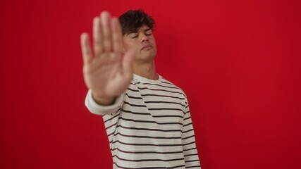 Canvas Print - Stop gesture by a young hispanic man wearing stripes t-shirt, standing with serious, negative warning expression and hand palm over isolated red background