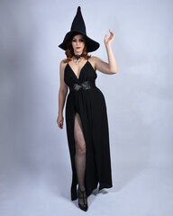 Wall Mural - Full length portrait of beautiful female red head model wearing  glamorous black witch dress with pointy hat halloween costume.   walking pose with gestural arms, isolated figure on studio background