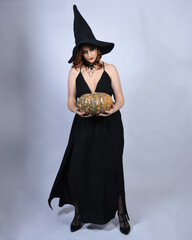 Canvas Print - portrait of beautiful female model wearing glamorous black witch dress, pointy hat halloween costume. walking pose holding pumpkin, isolated figure on studio background