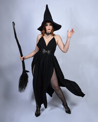 Poster - portrait of beautiful female model wearing glamorous black witch dress, pointy hat halloween costume. walking pose holding broomstick, isolated figure on studio background