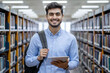 © Elathi - Cheerful Indian student in blue shirt with backpack and a tablet on hand in college library