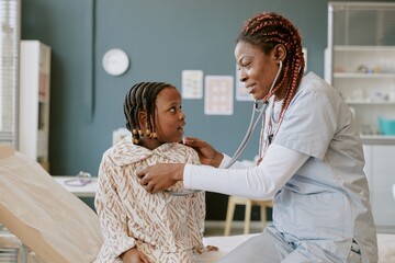 Medical professional with stethoscope examining young patient in brightly lit clinic setting. Child looking at doctor attentively while seated on examination table and nurse listening to heartbeat