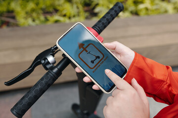 Hands holding smartphone displaying scooter sharing app interface by e-scooter handlebar. Person using technology for urban transportation solution