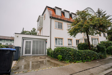 Munich, Germany - September 14, 2024 - Modern white house with large windows, a garage, and a small tree, on a rainy day, with wet pavement and green hedges surrounding the property..