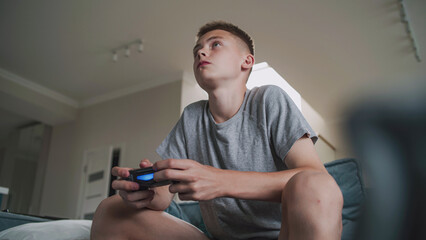 Concentrated Caucasian teenager plays online video game on console using wireless controller, sits on sofa in cozy living room. Teenage boy spending leisure time at home. Lifestyle concept. Low angle.