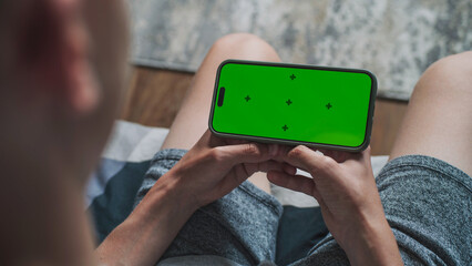 Poster - caucasian teenager holds mobile phone with green screen, watches video or movie sitting on sofa. you