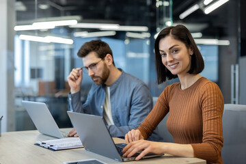 Wall Mural - Two business professionals collaborating in a modern office environment. Woman smiling while typing on laptop, man in background working on laptop with thoughtful expression