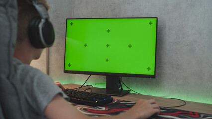 Poster - caucasian teenager wearing headphones looks at personal computer monitor with green screen. young bo