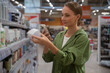 © Grustock - Young woman choosing cosmetics in a supermarket, comparing products and checking information on her smartphone. Engaging in informed decision-making while shopping for beauty items