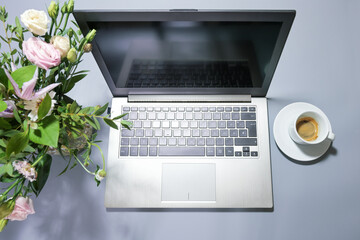 Silver colored laptop on a gray desk with a bouquet of flowers and a coffee cup, business workplace or home office, top view from above, copy space, selected focus